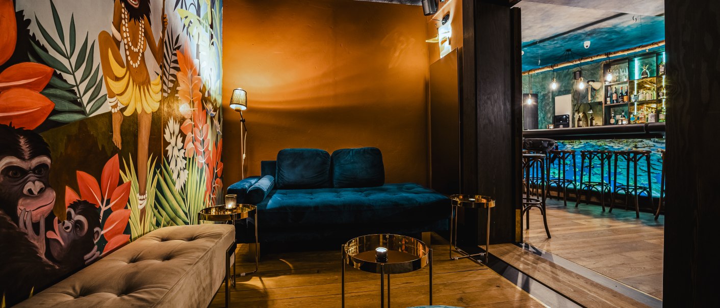 Stilvolle Bar mit blauen Sofas, bunter Wandmalerei und Holzboden. Im Hintergrund ist eine gut bestückte Theke zu sehen., © Holzmaler