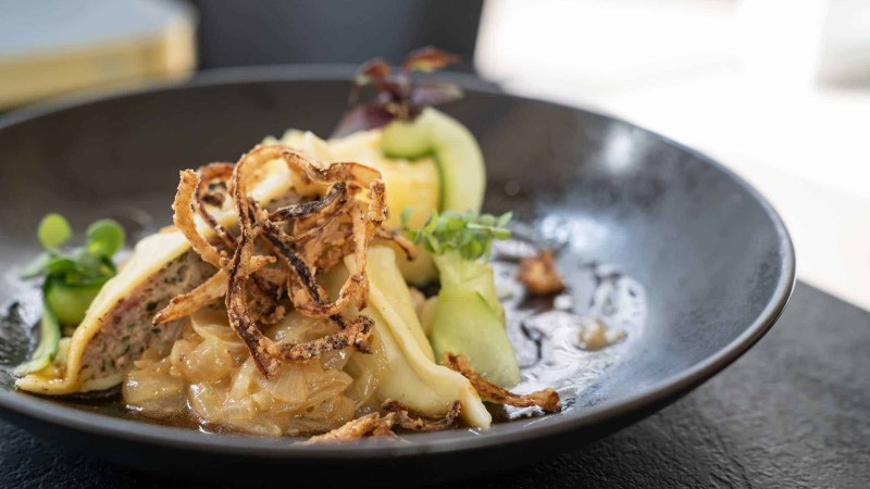 Ein elegantes Gericht mit gefüllten Nudeln, knusprigen Zwiebeln und frischen Kräutern auf einem schwarzen Teller angerichtet., © Flairhotel Vier Jahreszeiten Ein elegantes Gericht mit gefüllten Nudeln, knusprigen Zwiebeln und frischen Kräutern auf einem schwarzen Teller angerichtet., © Flairhotel Vier Jahreszeiten