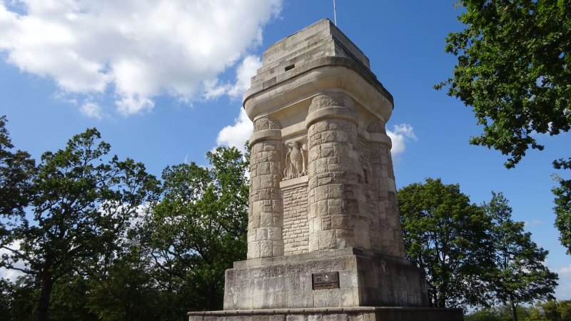Ein massiver Bismarckturm aus Stein steht auf einem Platz, umgeben von Bäumen und unter einem blauen Himmel mit wenigen Wolken., © SMG