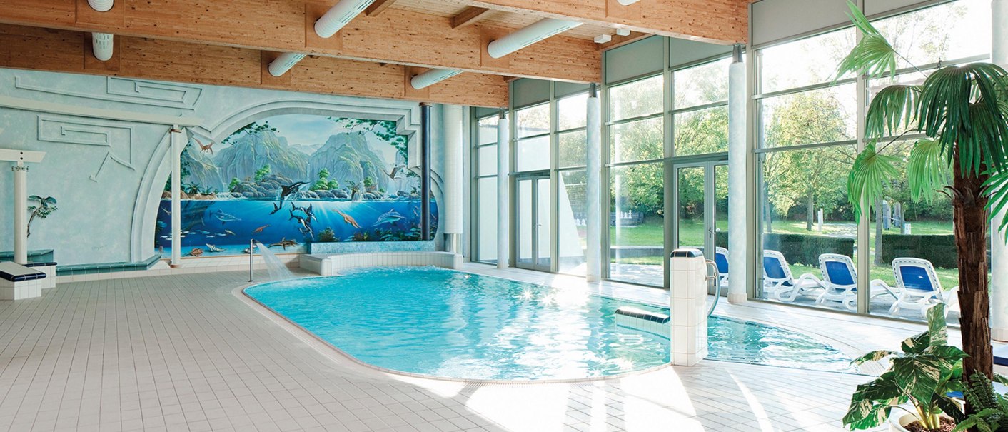 Ein Innenpool mit einem Wandgemälde von Meerestieren. Große Fenster lassen viel Licht herein. Liegestühle stehen am Rand, eine Palme im Vordergrund., © Seminaris Hotel Bad Boll