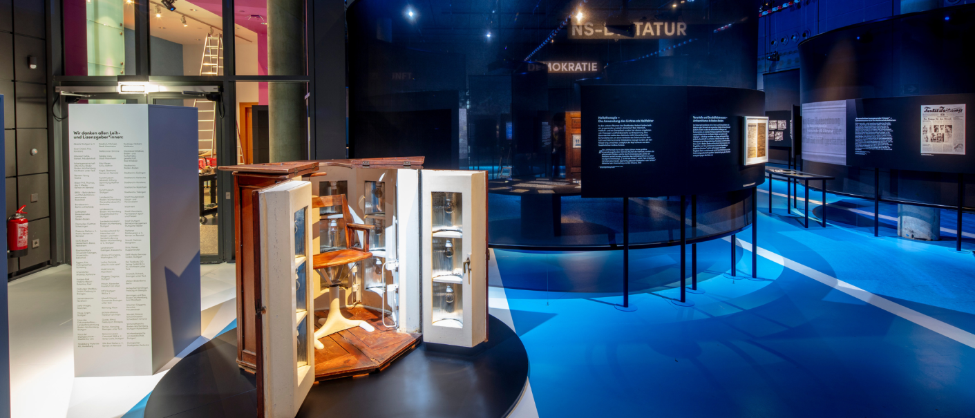 Ein moderner Ausstellungsraum mit blauen Böden, Texttafeln und einem geöffneten Holzschrank mit Stuhl. Im Hintergrund sind weitere Exponate zu sehen., © Frei_Schwimmen_Daniel_Stauch Ein moderner Ausstellungsraum mit blauen Böden, Texttafeln und einem geöffneten Holzschrank mit Stuhl. Im Hintergrund sind weitere Exponate zu sehen., © Frei_Schwimmen_Daniel_Stauch