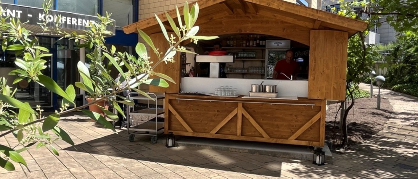 A wooden stand in front of a modern building with a glass fa&ccedil;ade. Plants can be seen in the foreground, partially covering the stand., &copy; TOMAS