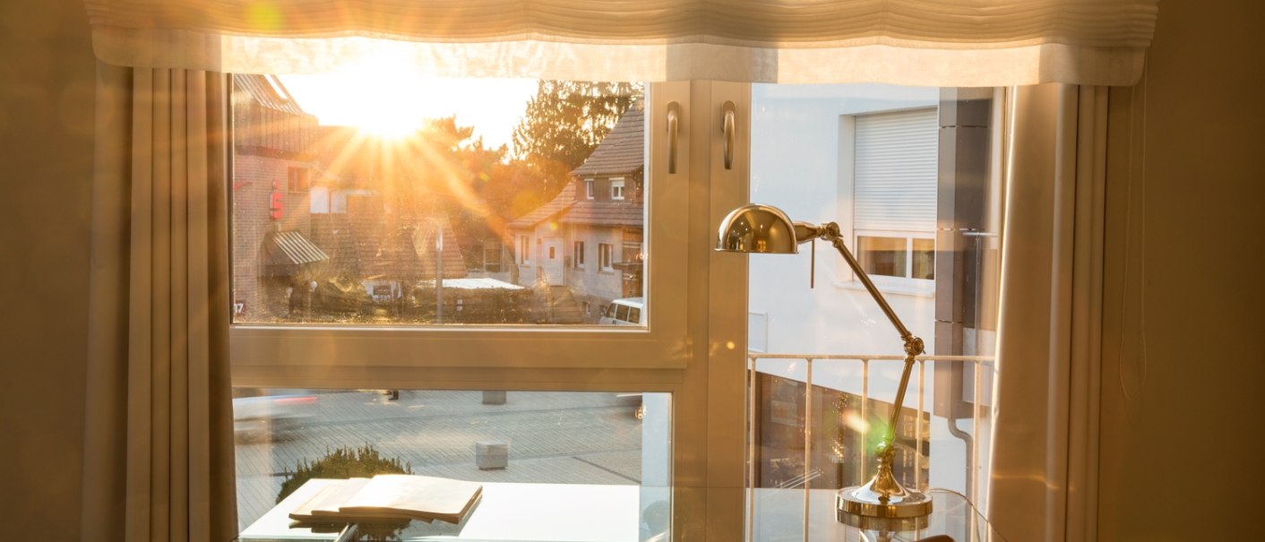 A modern desk with a lamp stands in front of a window. The sun shines in and illuminates the room warmly., © Hotel LOGIS A modern desk with a lamp stands in front of a window. The sun shines in and illuminates the room warmly., © Hotel LOGIS