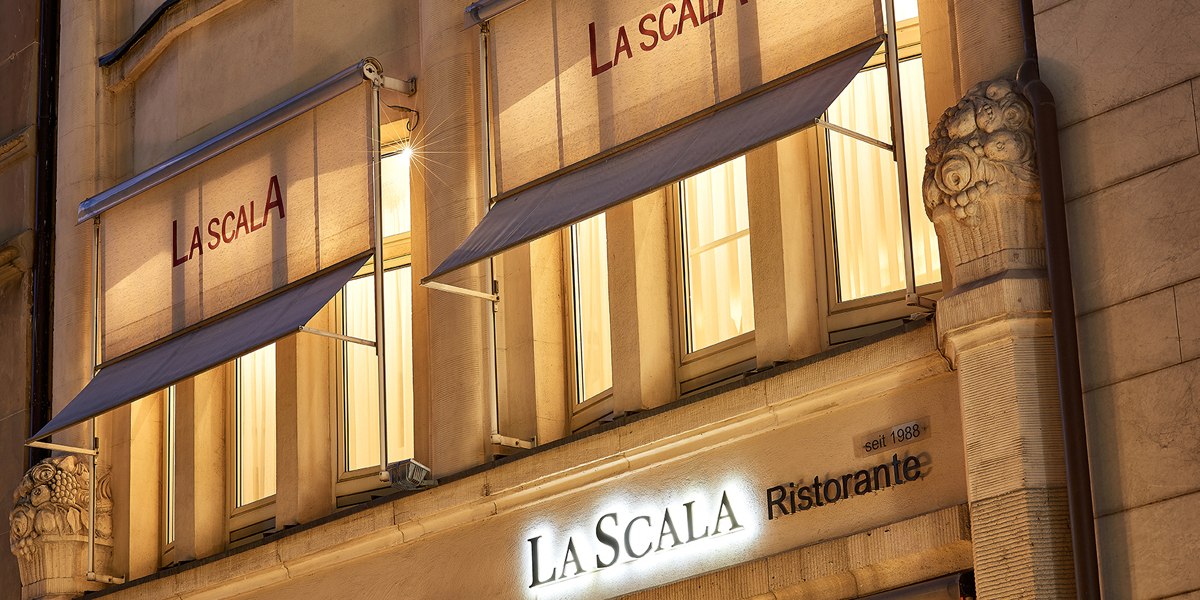 Illuminated façade of the La Scala restaurant in Stuttgart with awnings and decorative columns at night., © La Scala