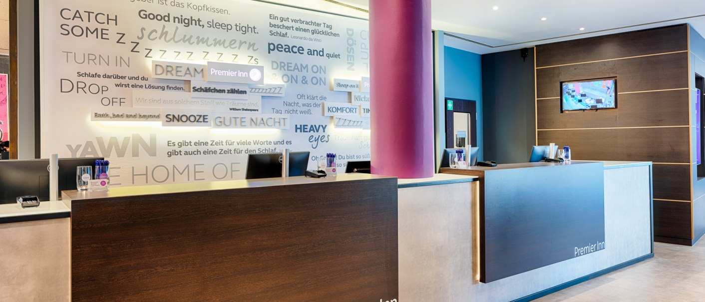 Modern hotel reception with two desks, purple pillar and wall full of bedroom quotes. Premier Inn logo visible., © Premier Inn Germany Modern hotel reception with two desks, purple pillar and wall full of bedroom quotes. Premier Inn logo visible., © Premier Inn Germany
