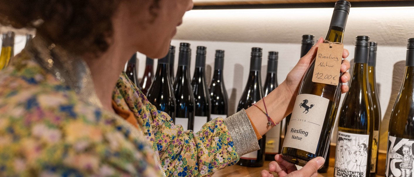 Eine Person betrachtet eine Weinflasche mit der Aufschrift 'Riesling Natur' in einer Weinbar. Im Hintergrund stehen weitere Weinflaschen., &copy; SMG, Martina Denker
