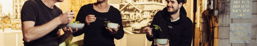 Three men sit together laughing and eating salad in a cozy, artistically designed room., &copy; Vhy! GmbH