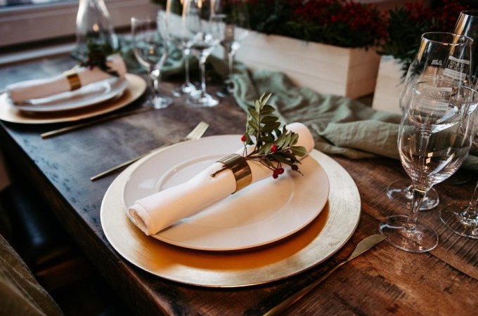 Elegante Tischdekoration mit goldenen Tellern, weißen Servietten und grünen Zweigen auf einem Holztisch, umgeben von Weingläsern., © Cool-Tours StattReisen Elegante Tischdekoration mit goldenen Tellern, weißen Servietten und grünen Zweigen auf einem Holztisch, umgeben von Weingläsern., © Cool-Tours StattReisen