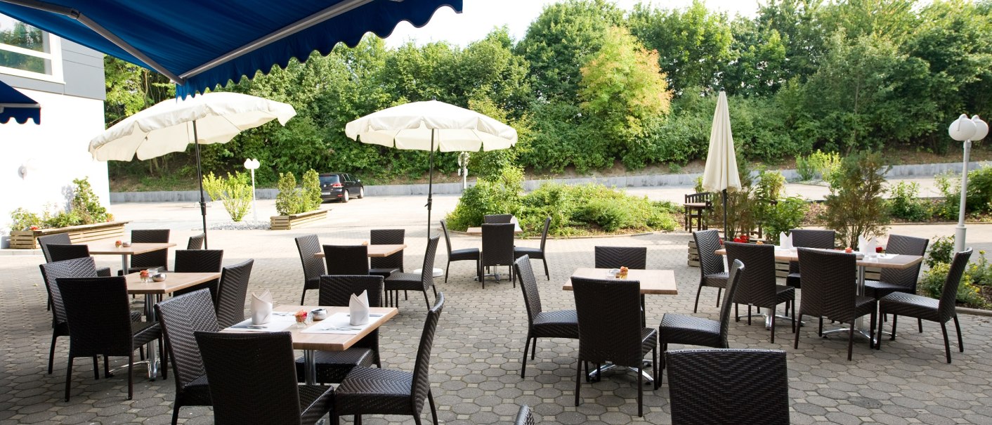 An outdoor terrace with empty tables and chairs, covered by blue awnings. White parasols and green bushes in the background., © Hotel Fortuna