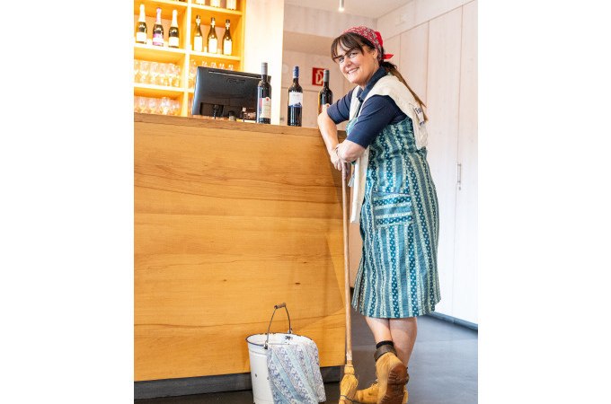 Frau in traditioneller Kleidung lehnt lächelnd an einer Theke mit Weinflaschen. Ein Eimer und Besen stehen daneben., © Stuttgart Marketing GmbH