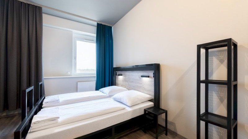 Modernes Doppelzimmer mit zwei Einzelbetten, einem kleinen Nachttisch und einem Regal. Helle W&auml;nde und Vorh&auml;nge in Blau und Grau., &copy; A&O Hostels Marketing GmbH