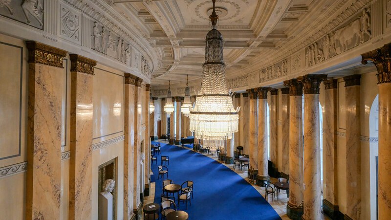 Elegant hall with blue carpets, marble columns and large chandeliers hanging from the ceiling. Tables and chairs are arranged along the carpet., &copy; W&uuml;rttembergische Staatstheater Stuttgart