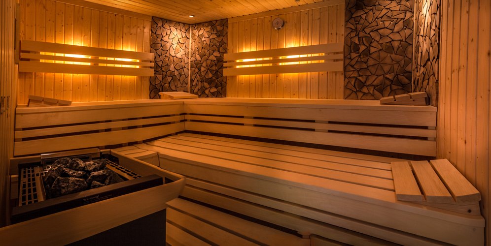 A modern sauna with wooden benches, warm lighting, and a stone-lined stove. The walls are made of wood and decorative stone., &copy; Kulinarium an der Glems