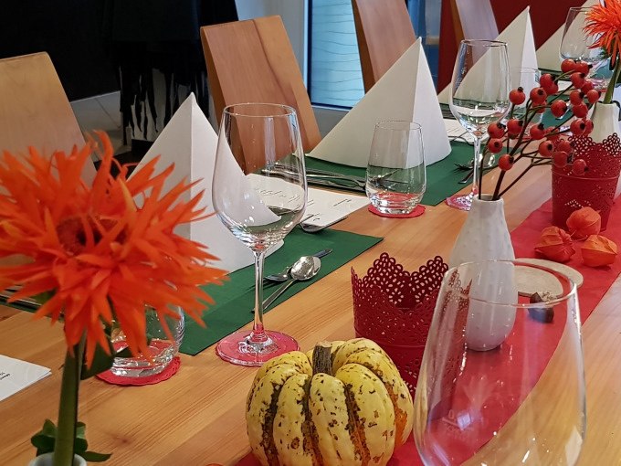 Ein festlich gedeckter Tisch mit Gläsern, weißen Servietten, orangefarbenen Blumen und herbstlicher Dekoration in einem modernen Raum., © Kultweine-Kochwelten-Kunstücke - Kochschule Sonja Lenz Ein festlich gedeckter Tisch mit Gläsern, weißen Servietten, orangefarbenen Blumen und herbstlicher Dekoration in einem modernen Raum., © Kultweine-Kochwelten-Kunstücke - Kochschule Sonja Lenz