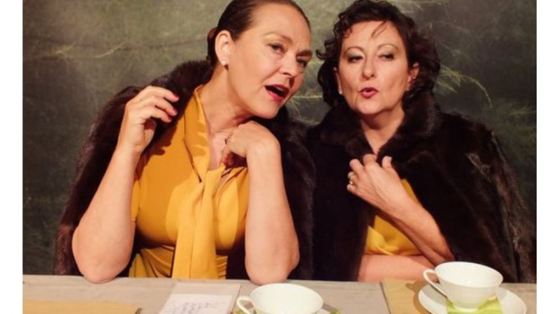 Zwei Frauen in gelber Kleidung und Pelzstolen sitzen an einem Tisch mit Teetassen und scheinen zu flüstern., © Fotocredit Theater La Lune Stuttgart Zwei Frauen in gelber Kleidung und Pelzstolen sitzen an einem Tisch mit Teetassen und scheinen zu flüstern., © Fotocredit Theater La Lune Stuttgart