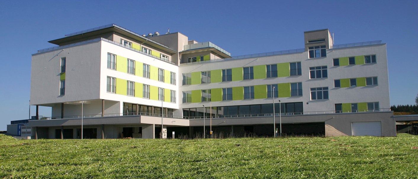 Modern hotel building with green accents, surrounded by a meadow, under a clear blue sky., © TOMAS