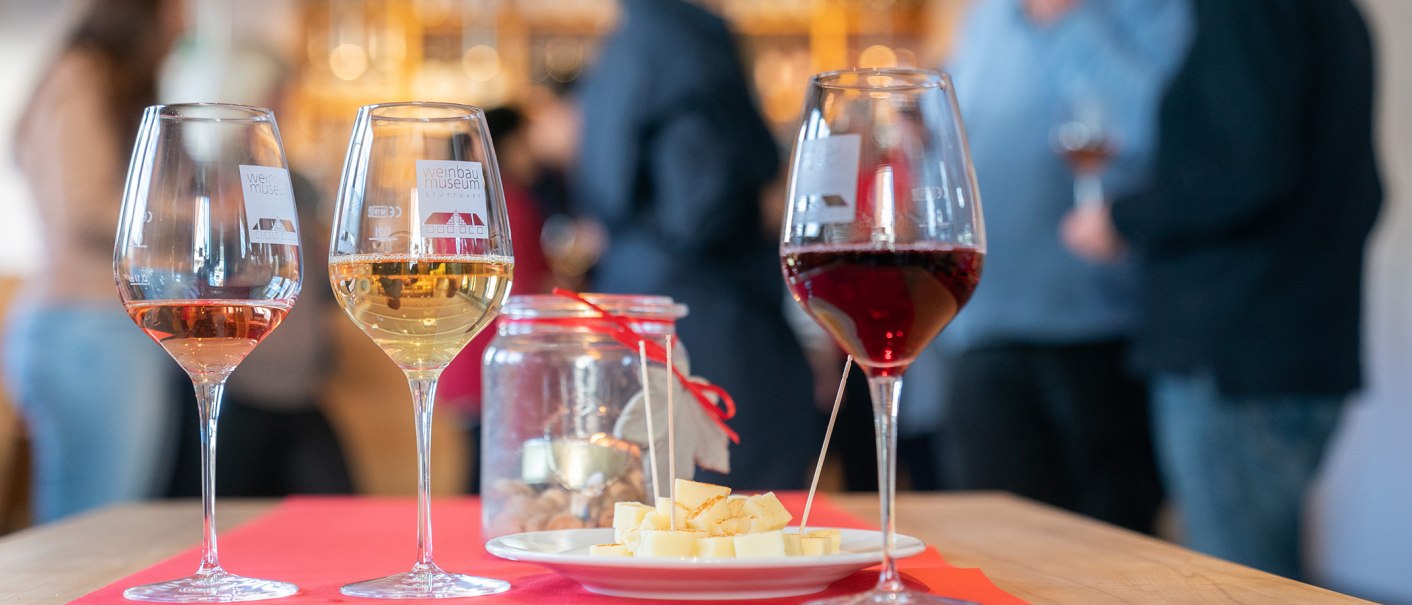 Three wine glasses with red, white and rosé wine stand on a table. Next to them is a plate with cheese cubes and a glass with a candle. People in the background., © Thomas Niedermüller Three wine glasses with red, white and rosé wine stand on a table. Next to them is a plate with cheese cubes and a glass with a candle. People in the background., © Thomas Niedermüller