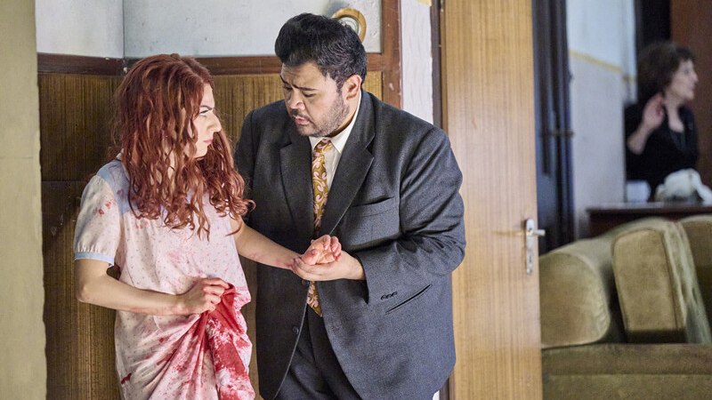 Ein Mann im Anzug h&auml;lt eine Frau in einem blutbefleckten Kleid. Im Hintergrund ist eine weitere Person zu sehen., &copy; W&uuml;rttembergische Staatstheater Stuttgart