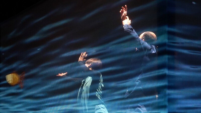 Zwei Personen tanzen vor einer Projektion von Wellen, die eine Meereslandschaft darstellen. Die Szene wirkt künstlerisch und dynamisch., © FITZ Das Theater animierter Formen Zwei Personen tanzen vor einer Projektion von Wellen, die eine Meereslandschaft darstellen. Die Szene wirkt künstlerisch und dynamisch., © FITZ Das Theater animierter Formen