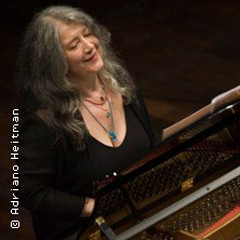 Eine Frau mit grauem Haar sitzt l&auml;chelnd am Klavier auf einer B&uuml;hne. Sie tr&auml;gt ein schwarzes Oberteil und eine Halskette. Der Raum ist dunkel beleuchtet., &copy; links im Bild