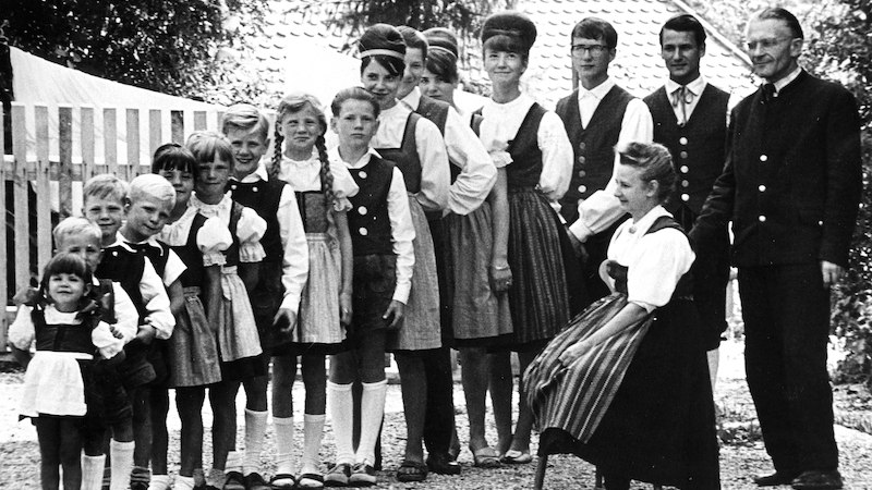 Eine große Familie in traditioneller Kleidung posiert vor einem Haus. Die Frauen tragen Dirndl, die Männer Anzüge. Es ist ein Schwarz-Weiß-Foto., © Theaterhaus Stuttgart e.V. Eine große Familie in traditioneller Kleidung posiert vor einem Haus. Die Frauen tragen Dirndl, die Männer Anzüge. Es ist ein Schwarz-Weiß-Foto., © Theaterhaus Stuttgart e.V.