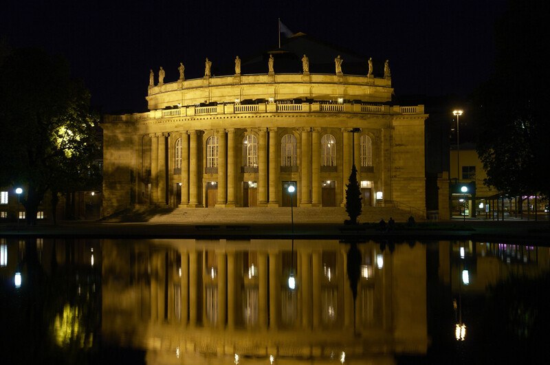 Beleuchtetes historisches Gebäude mit Säulen, nachts aufgenommen. Die Fassade spiegelt sich im Wasser davor wider., © Württembergische Staatstheater Stuttgart