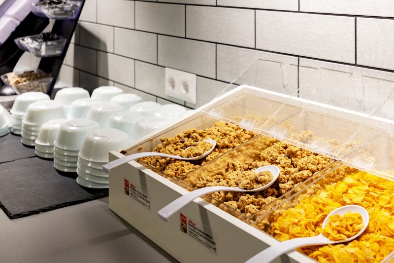 Breakfast buffet with different types of muesli and cornflakes in containers, white bowls stacked next to them., &copy; A&O Hostels Marketing GmbH