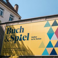 Gelbes Schild mit 'Buch & Spiel', bunten Dreiecken und Betonwand im Hintergrund. Kultur in St. Stefan, geöffnet Mo-Sa 10-18 Uhr., © SMG, Sarah Schmid Gelbes Schild mit 'Buch & Spiel', bunten Dreiecken und Betonwand im Hintergrund. Kultur in St. Stefan, geöffnet Mo-Sa 10-18 Uhr., © SMG, Sarah Schmid