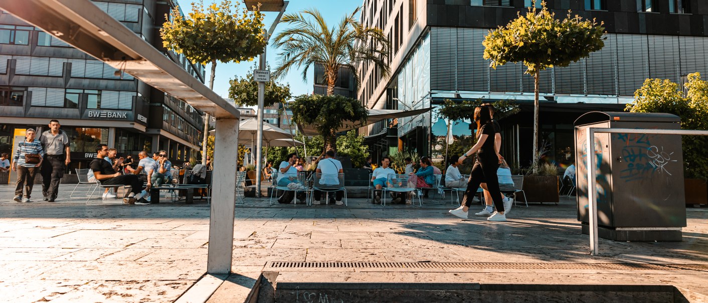 Menschen sitzen in einem Straßencafé vor modernen Gebäuden. Die Sonne scheint, und Bäume sowie eine Palme schmücken den Platz., © SMG, Sarah Schmid
