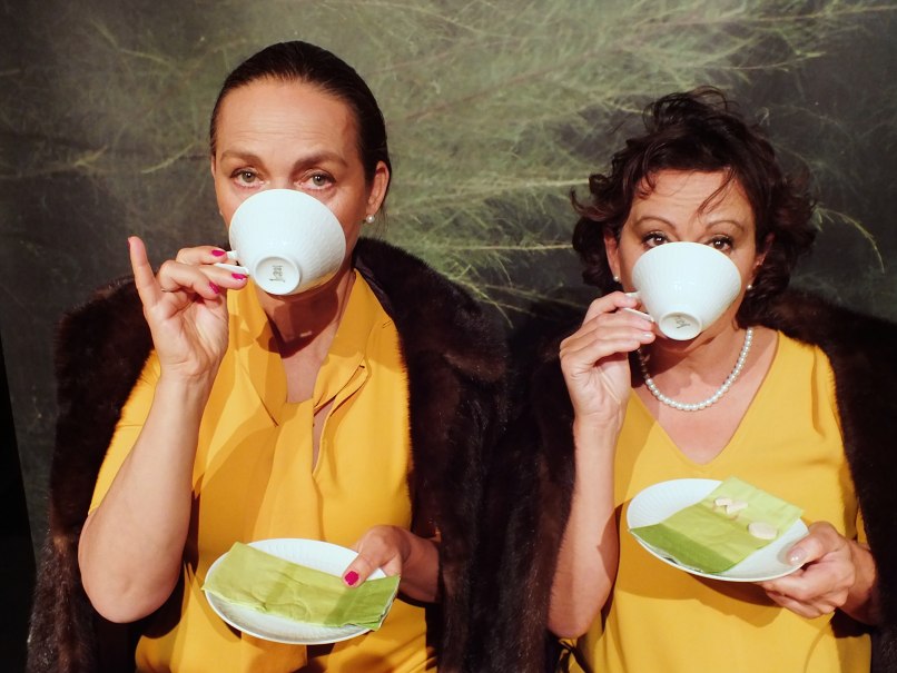 Zwei Frauen in gelber Kleidung trinken Tee aus weißen Tassen. Sie halten Untertassen mit grünen Servietten und Keksen., © Dieter Nelle