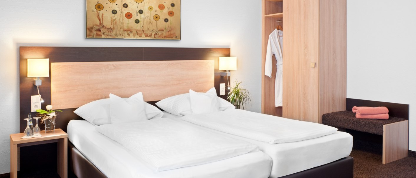 A modern hotel room with a double bed, nightstand, closet, and wall hanging. A bathrobe hangs in the closet, and towels are laid out on a bench., &copy; TOMAS