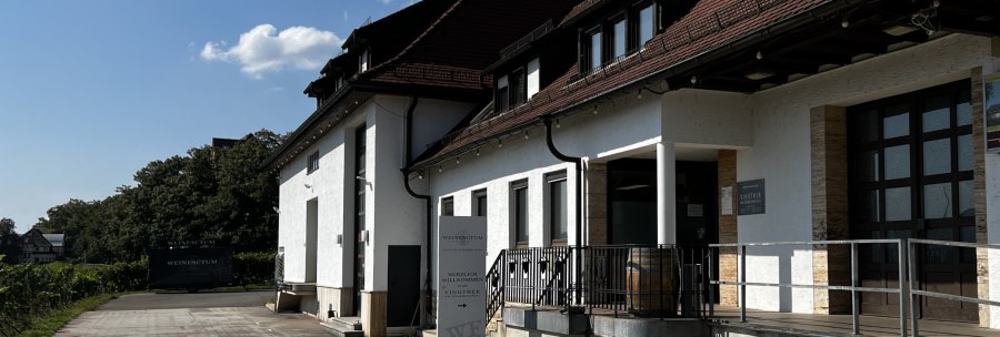 White building with red roof, vinotheque sign and wine barrel in front of the entrance. Surrounded by trees and a clear sky., &copy; Weinfactum Bad Cannstatt