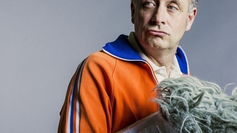 Mann in orangefarbenem Trainingsanzug mit blauem Kragen, h&auml;lt Mikrofon mit Windschutz. Hintergrund grau., &copy; Theaterhaus Stuttgart e.V.