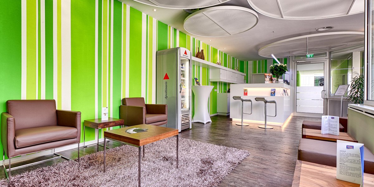 Moderne Rezeption mit grüner gestreifter Wand, braunen Sesseln, Teppich und weißem Tresen. Helle, einladende Atmosphäre mit Pflanzen und Broschüren., © attimo Moderne Rezeption mit grüner gestreifter Wand, braunen Sesseln, Teppich und weißem Tresen. Helle, einladende Atmosphäre mit Pflanzen und Broschüren., © attimo