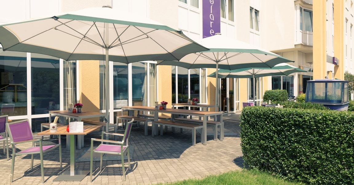 A terrace with wooden tables, purple chairs, and large umbrellas in front of a yellow building with large windows., &copy; Mercure Hotel Stuttgart Gerlingen