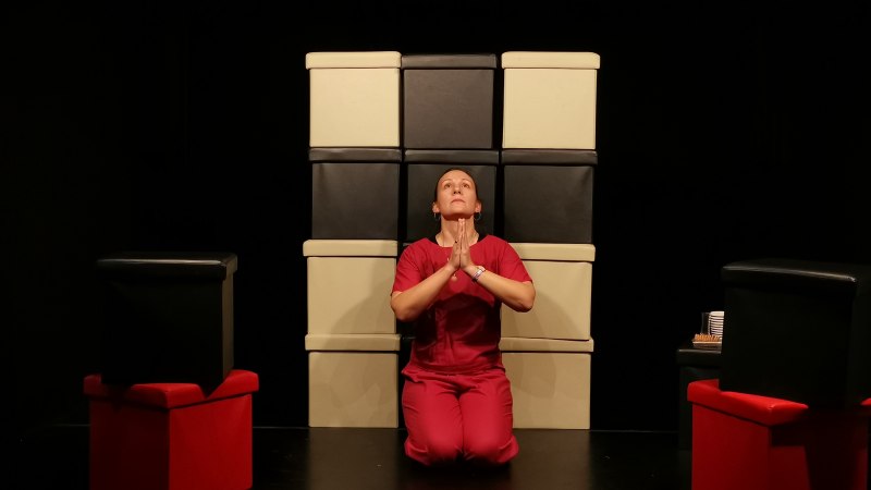 Person in roter Kleidung kniet vor gestapelten Boxen, Hände betend gefaltet. Dunkler Hintergrund, rote und schwarze Boxen., © Maja Kles Person in roter Kleidung kniet vor gestapelten Boxen, Hände betend gefaltet. Dunkler Hintergrund, rote und schwarze Boxen., © Maja Kles