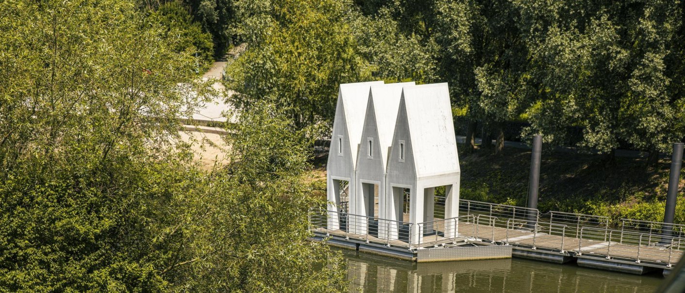 A modern bathhouse with pointed roofs stands on a river, surrounded by lush greenery. It is accessible via a footbridge., &copy; SMG Stuttgart Marketing GmbH - Sarah Schmid