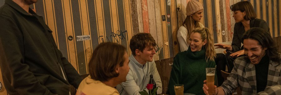 Eine Gruppe von Menschen sitzt in einer gemütlichen Bar und lacht. Sie genießen Getränke und unterhalten sich lebhaft. Der Raum hat eine warme Atmosphäre., © SMG, Martina Denker