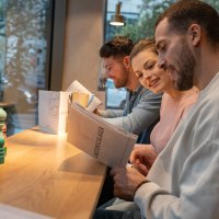 Three people are sitting at a table in a restaurant reading the menu. In the background is a window with a view outside., © Stuttgart-Marketing GmbH, Martina Denker Three people are sitting at a table in a restaurant reading the menu. In the background is a window with a view outside., © Stuttgart-Marketing GmbH, Martina Denker