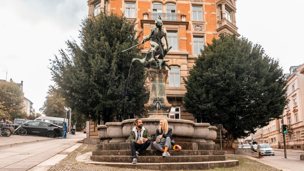 Two people sit on the steps of a fountain in an urban setting, surrounded by historic buildings and trees., &copy; SMG, Sarah Schmid