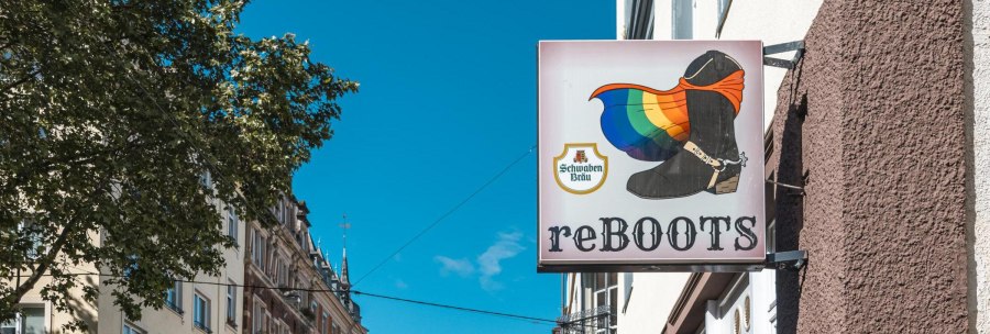 A sign with a boot and a rainbow flag, next to it the inscription 'reBOOTS'. Buildings and a tree can be seen in the background., © SMG Stuttgart Marketing GmbH - Sarah Schmid A sign with a boot and a rainbow flag, next to it the inscription 'reBOOTS'. Buildings and a tree can be seen in the background., © SMG Stuttgart Marketing GmbH - Sarah Schmid