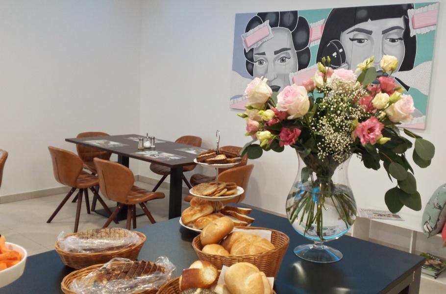 Ein Frühstücksbüffet mit Brötchen, Gebäck und einem Blumenstrauß. Im Hintergrund ein Tisch mit Stühlen und ein Kunstwerk an der Wand., © TOMAS Ein Frühstücksbüffet mit Brötchen, Gebäck und einem Blumenstrauß. Im Hintergrund ein Tisch mit Stühlen und ein Kunstwerk an der Wand., © TOMAS