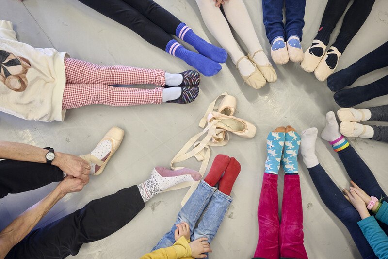 Children and adults sit in a circle on the floor, their legs stretched out. Various colorful socks and ballet shoes can be seen., &copy; W&uuml;rttembergische Staatstheater Stuttgart