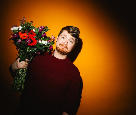 A person holds a colorful bouquet of flowers in front of a bright orange background., &copy; Rosenau Kultur e.V.