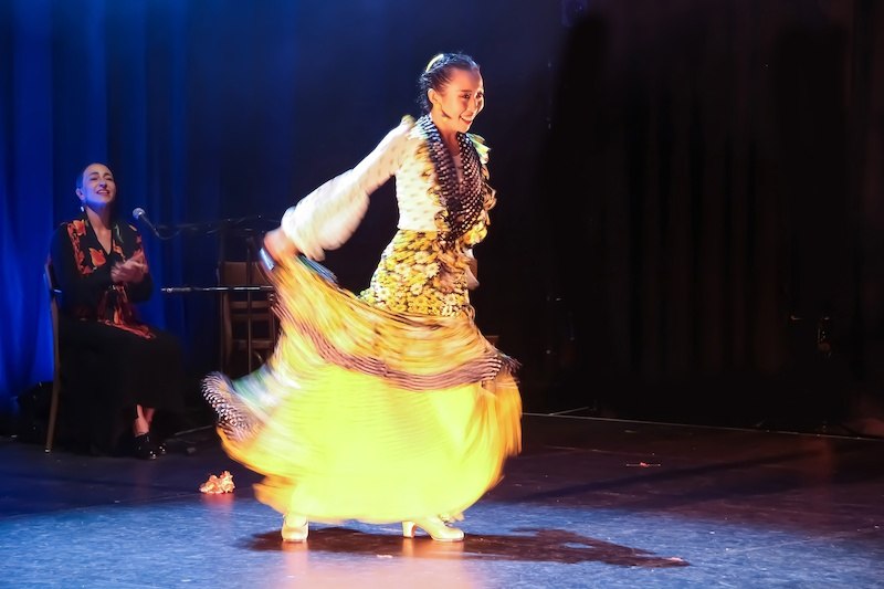 Eine Flamenco-T&auml;nzerin in einem leuchtend gelben Rock tanzt auf der B&uuml;hne, w&auml;hrend eine Musikerin im Hintergrund applaudiert., &copy; Stuttgarter Flamenco Festival gUG