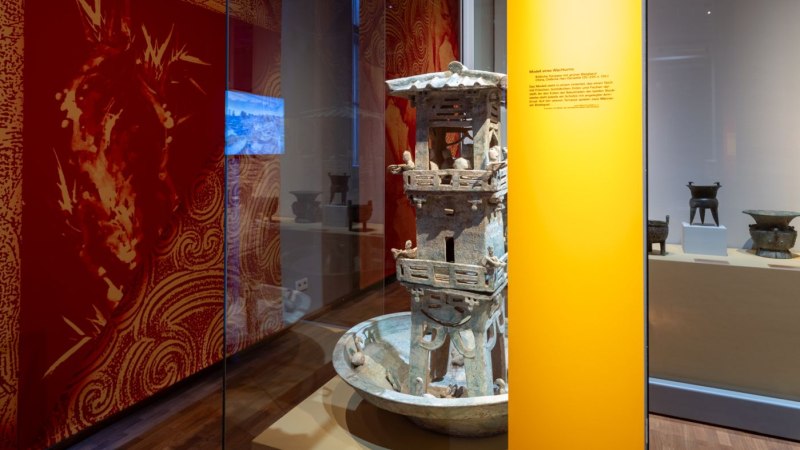 Ein Ausstellungsraum zeigt ein Modell eines chinesischen Wachtturms und antike Gefäße. Die Wände sind mit roten Mustern dekoriert., © Linden-Museum Stuttgart