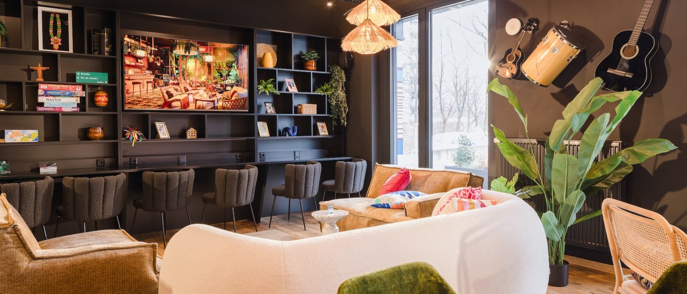 Ein gemütlicher Raum mit Sofas, Regalen voller Bücher und Spiele, Musikinstrumenten an der Wand und großen Fenstern. Pflanzen und warme Beleuchtung schaffen eine einladende Atmosphäre., © TOMAS