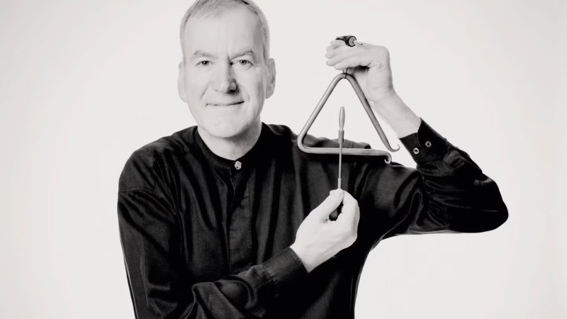 Ein Mann in schwarzer Kleidung hält ein Triangel und einen Schlägel in den Händen, lächelnd in die Kamera schauend., © Marco Borggreve Ein Mann in schwarzer Kleidung hält ein Triangel und einen Schlägel in den Händen, lächelnd in die Kamera schauend., © Marco Borggreve