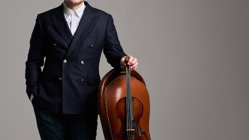 Ein Mann in elegantem Anzug lehnt an einem braunen Lederstuhl, auf dem eine Geige liegt. Der Hintergrund ist neutral grau., © Klassische Philharmonie Bonn e.V.