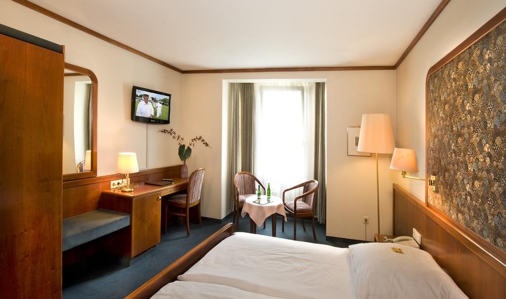 Gemütliches Hotelzimmer mit Doppelbett, Fernseher, Schreibtisch und Sitzbereich vor einem Fenster. Holzmöbel und warme Beleuchtung dominieren den Raum., © Hotel am Schelztor Gemütliches Hotelzimmer mit Doppelbett, Fernseher, Schreibtisch und Sitzbereich vor einem Fenster. Holzmöbel und warme Beleuchtung dominieren den Raum., © Hotel am Schelztor